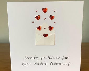 May include: A white greeting card with a paper envelope and red heart-shaped embellishments. The card reads, "Sending you love on your Ruby Wedding Anniversary..."