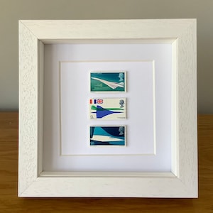 May include: A white framed artwork displaying three postage stamps depicting the Concorde supersonic jet. The stamps feature blue, green, and white designs, with the word "Concorde" printed on two of them.