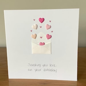 Puede incluir: Una tarjeta blanca con un sobre blanco y corazones rosas. El texto en la tarjeta dice "Sending you love on your birthday."