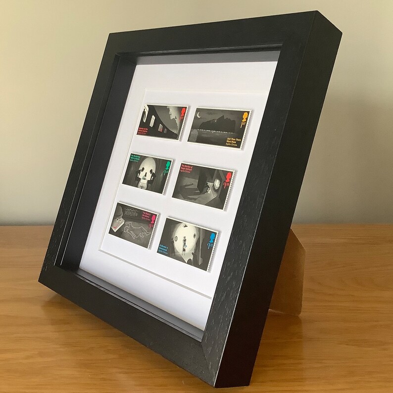 May include: A black framed display with a white mat showcasing a collection of six postage stamps. The stamps feature black and white illustrations with colorful accents. The stamps are arranged in two rows of three.