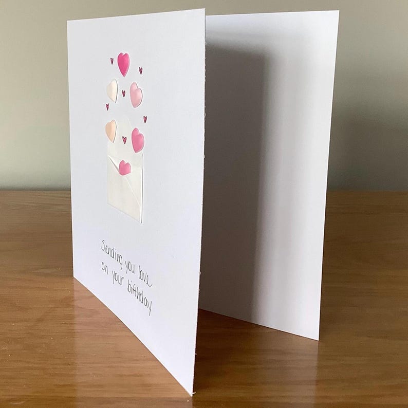 Puede incluir: Una tarjeta blanca con un sobre rosa y corazones rosas. El texto en la tarjeta dice "Sending you love on your birthday."