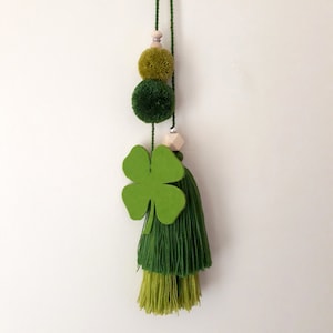 Może przedstawiać: Zielony urok w kształcie koniczyny z zielonymi pomponami i zielonym frędzlem wiszącym na sznurku.