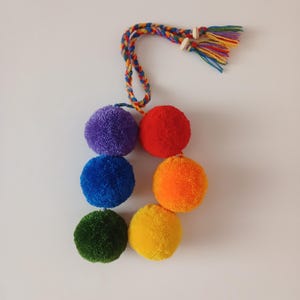 May include: A handmade rainbow pom-pom garland with six colorful pom-poms in purple, red, blue, orange, green, and yellow. The garland features a braided cord and wooden beads at the top, with a tassel of colorful threads.