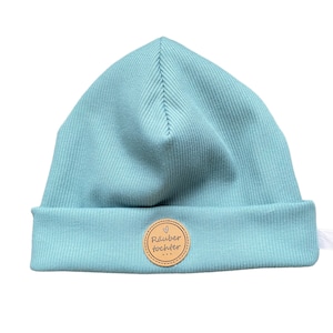 Könnte beinhalten: Hellblaues Rippstrick-Beanie mit umgeschlagenem Saum. Ein kleines, rundes, braunes Lederpatch mit dem Text "Räuber Tochter" ist vorne aufgenäht. Die Mütze ist aus einem weichen, dehnbaren Material gefertigt.