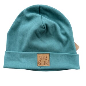 Peut inclure: Béret bleu sarcelle avec un patch en cuir marron qui dit "Wild Child" en cursive.