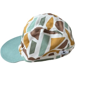 May include: A cap with a light blue brim and a patterned crown. The crown features a white background with abstract shapes in brown, yellow, and light green. The cap has a small logo on the side.