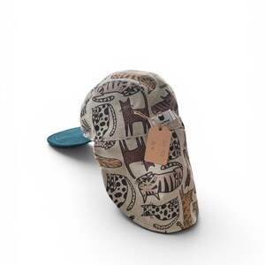 May include: A beige baseball cap featuring a repeating pattern of various brown and black cats in different poses. The cap has a teal underside on the brim and a small brown paper tag attached with twine, displaying the text SA-53 and 32 €.