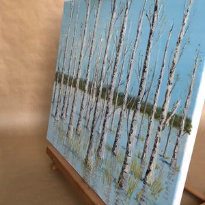 May include: A painting on canvas depicting a birch tree forest with white trunks and branches. The trees are reflected in the blue water below. The painting is on an easel.