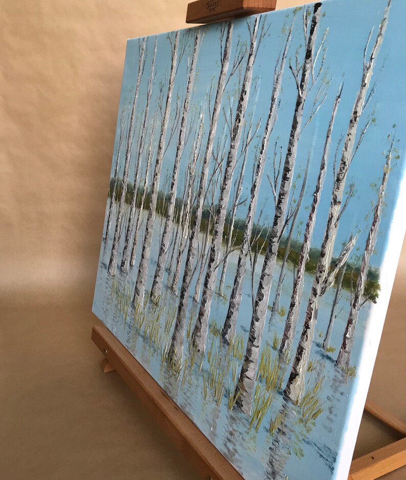 May include: A painting on canvas depicting a birch tree forest with white trunks and branches. The trees are reflected in the blue water below. The painting is on an easel.