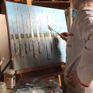 May include: A person in a white coat is painting a canvas with a palette knife. The canvas depicts a winter scene with a row of birch trees. The artist is using a palette knife to apply white paint to the canvas. The artist has a palette of paint colors in front of them.