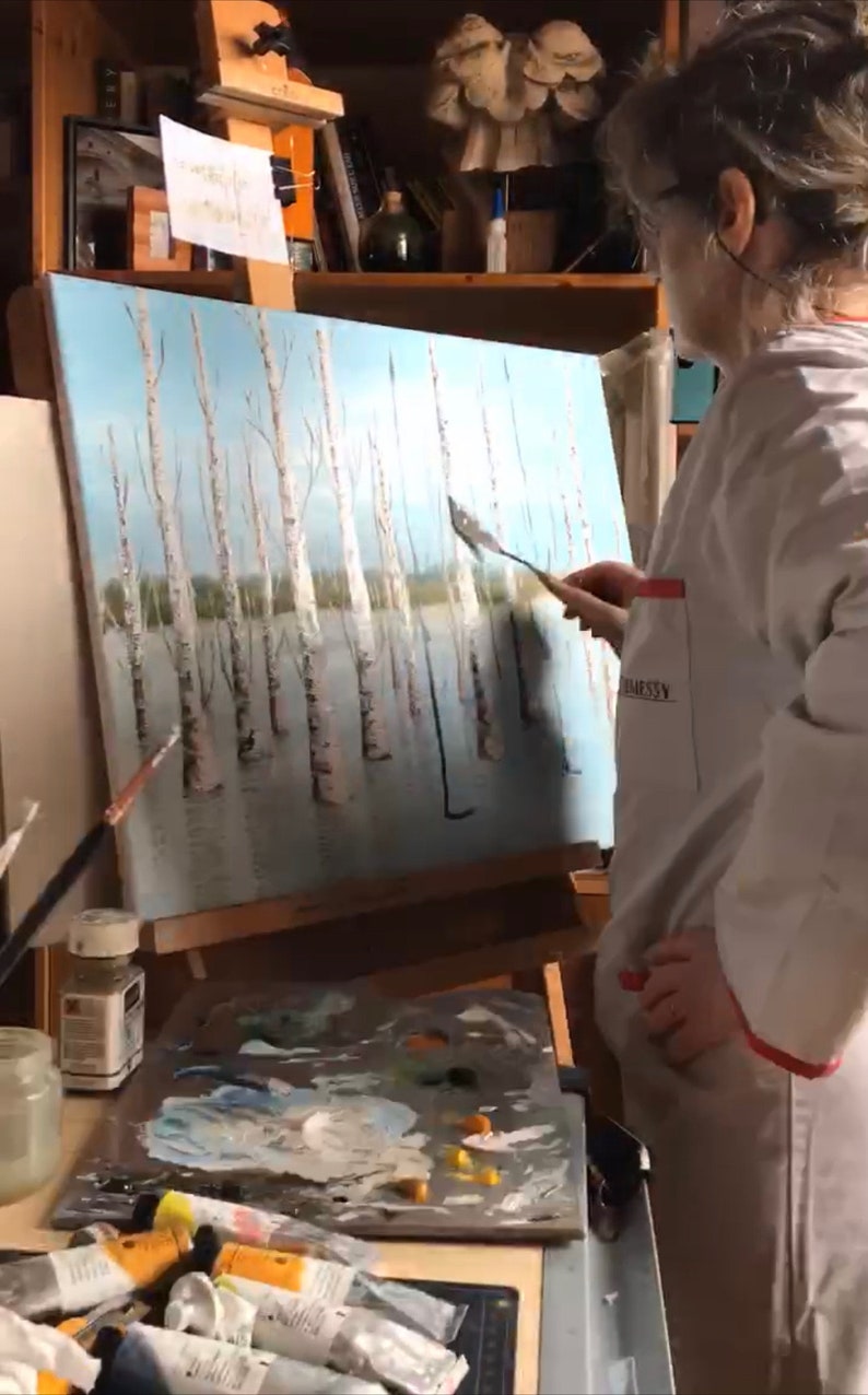 May include: A person in a white coat is painting a canvas with a palette knife. The canvas depicts a winter scene with a row of birch trees. The artist is using a palette knife to apply white paint to the canvas. The artist has a palette of paint colors in front of them.