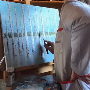 May include: A person paints a canvas with a palette knife. The canvas depicts a landscape with trees and water. The person is wearing a white smock with a red pocket. The canvas is on an easel. There are paint tubes and a palette on the table.
