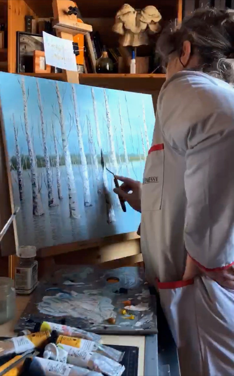May include: A person paints a canvas with a palette knife. The canvas depicts a landscape with trees and water. The person is wearing a white smock with a red pocket. The canvas is on an easel. There are paint tubes and a palette on the table.