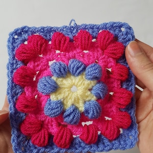 May include: A square, handmade crochet piece featuring a floral design. The piece has a blue border, with pink and magenta flower petals surrounding a yellow center. The crochet work is held in a person's hands.