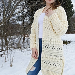May include: A white crocheted cardigan with a textured stitch pattern and bobble accents. The cardigan is open front and has long sleeves. The model is wearing the cardigan over a white t-shirt and blue jeans. The background is a snowy landscape with bare trees.