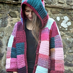 Könnte beinhalten: Eine Kapuzenjacke mit Patchwork-Muster in den Farben Rosa, Türkis und Braun. Die Jacke ist gehäkelt und hat einen Knopfverschluss.
