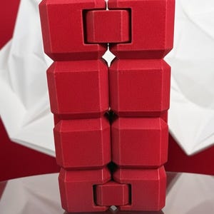 May include: A vibrant red, geometric sculpture composed of stacked, block-like forms. The sculpture has a textured surface and is displayed on a glass table. The design features interlocking components, creating a unique visual appeal.