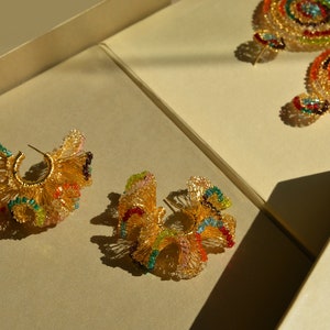 May include: A pair of gold hoop earrings with colorful beaded designs. The earrings are displayed on a beige surface.