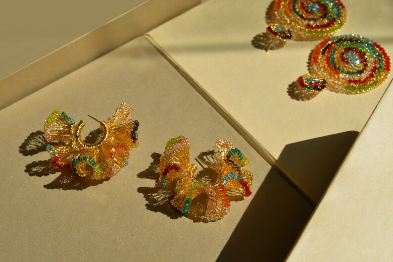 May include: A pair of gold hoop earrings with colorful beaded designs. The earrings are displayed on a beige surface.