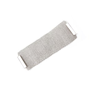 Puede incluir: Un brazalete plateado con un diseño de malla tejida. El brazalete tiene forma rectangular con dos barras plateadas en cada extremo. Se muestra sobre un fondo blanco.