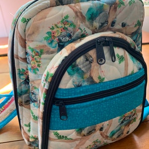 May include: A small quilted backpack with a koala print. The backpack has a blue and turquoise fabric panel with two zippered pockets. The backpack has a black zipper and black trim.