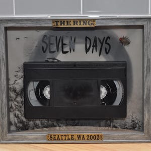 Peut inclure: Œuvre encadrée avec une cassette VHS noire, le texte "SEVEN DAYS", "THE RING" et "SEATTLE, WA 2002". Le cadre est gris avec des accents dorés.