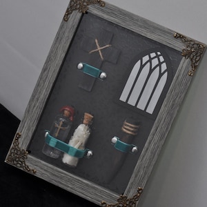 May include: Framed artwork with a dark background, a cross, a church window, and three small bottles. The bottles are secured with teal bands. The grey frame has decorative corner accents.