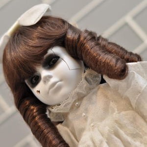 May include: A porcelain doll with brown curly hair and a white bonnet. The doll has a pale face with dark eye makeup and cracks. It wears a white dress with lace trim and pearl buttons, creating a spooky aesthetic.