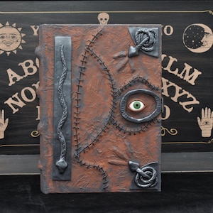 Peut inclure: Un livre marron texturé avec un design cousu et un grand œil vert au centre. La couverture présente un serpent argenté et deux accents argentés en forme de serpent. Le livre est posé sur un fond noir avec une planche Ouija.