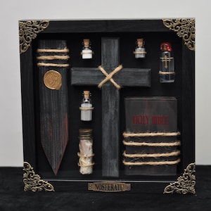 May include: A black shadow box frame with ornate gold corner accents containing a wooden cross, a wooden stake with a gold wax seal, a small book titled Holy Bible, and several glass vials filled with various substances, labeled Nosferatu.