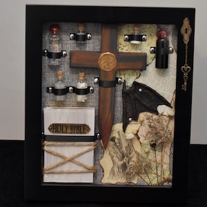 May include: A black shadow box frame containing a wooden cross with a gold wax seal, a white book labeled HOLY BIBLE wrapped in twine, small glass vials, a black bat wing, vintage map fragments, dried plants, and a hanging brass key charm.