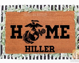May include: A rectangular brown coir doormat featuring the black text HOME with the United States Marine Corps emblem replacing the letter O, and the name HILLER printed below in black. The text ORCHID LANE BOUTIQUE appears at the top.