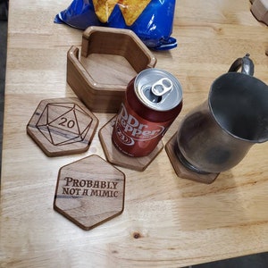 May include: Wooden coasters with laser-engraved designs. One coaster features a 20-sided die, another says "Probably not a mimic", and a third is a hexagon shape. A can of Dr Pepper and a silver mug are on top of the coasters.