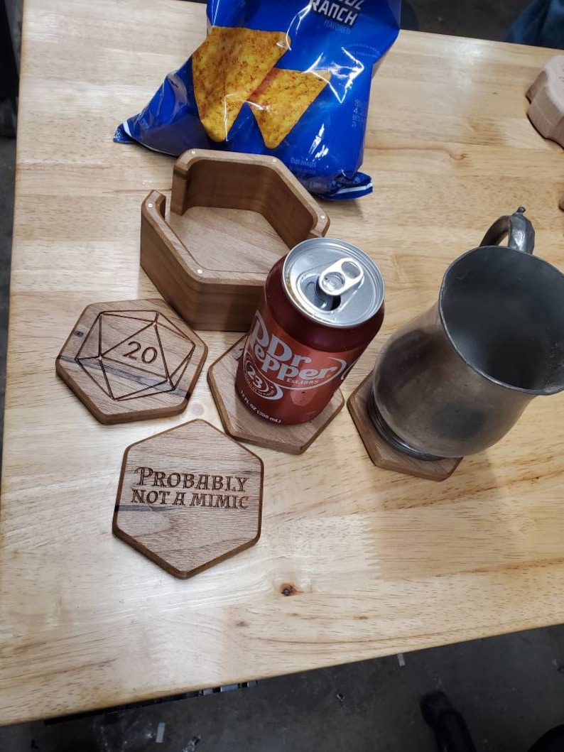 May include: Wooden coasters with laser-engraved designs. One coaster features a 20-sided die, another says "Probably not a mimic", and a third is a hexagon shape. A can of Dr Pepper and a silver mug are on top of the coasters.