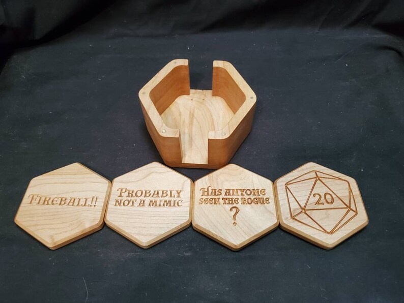 May include: Wooden box with a hinged lid and four hexagonal coasters. The coasters have engraved text: "Fireball!", "Probably not a mimic", "Has anyone seen the rogue?", and a 20-sided die with the number 20.
