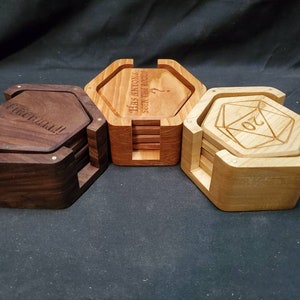 May include: Three sets of hexagonal wooden coasters. The coasters are stacked and have a wooden frame. The top coaster in each set has a different design: one says "The only thing seen the rocks", another says "The Cat's Meow", and the third has a 20-sided die design.