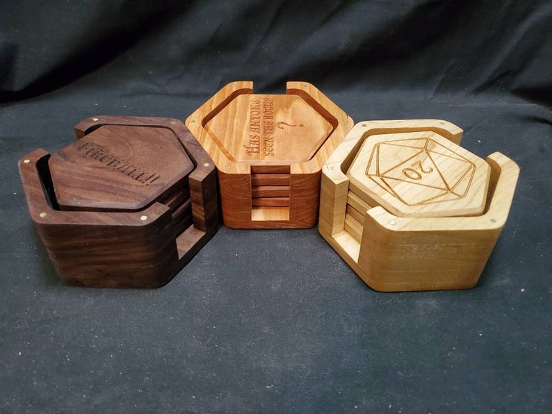 May include: Three sets of hexagonal wooden coasters. The coasters are stacked and have a wooden frame. The top coaster in each set has a different design: one says "The only thing seen the rocks", another says "The Cat's Meow", and the third has a 20-sided die design.