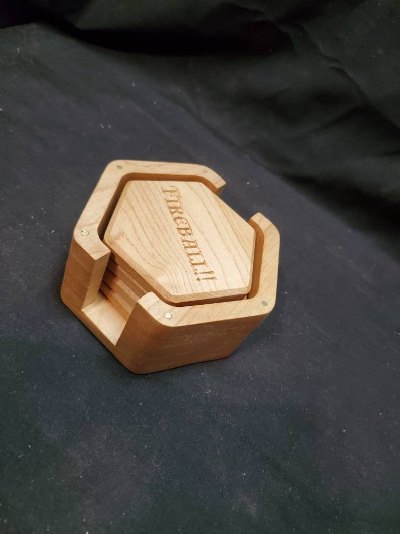 May include: Set of six hexagonal wooden coasters in a holder. The coasters are made of light wood and have the word "FIREBALL" engraved on the top. The holder is made of dark wood and has a hexagonal shape.