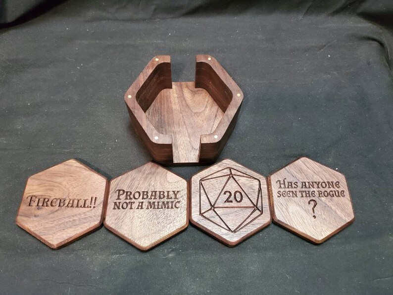 May include: Set of six wooden coasters with engraved designs. The coasters feature phrases like "Fireball!!", "Probably not a mimic", and "Has anyone seen the rogue?" One coaster has a 20-sided die design with the number 20 engraved on it. The coasters are made of dark wood and have a hexagonal shape.