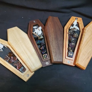 May include: Five wooden coffin-shaped boxes with lids open, revealing dice and miniature figures inside. The boxes are made of different types of wood, including walnut, cherry, and maple.