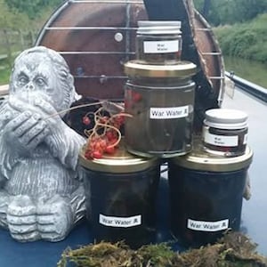 May include: Four glass jars with black lids and labels that say "War Water" in white text. The jars are arranged in a pyramid shape on a blue surface. There is a grey stone sculpture of a monkey in the background.