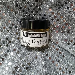 Può includere: Un piccolo barattolo di vetro con un coperchio nero e un'etichetta bianca che recita "The Enchanted Goddess Flying Ointment". Il barattolo è appoggiato su un tessuto argentato scintillante.