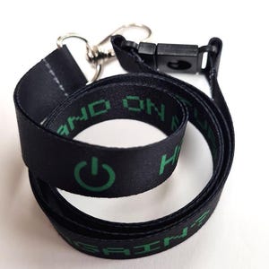 Könnte beinhalten: Schwarzes Schlüsselband mit grünem Text und einem Power-Button-Symbol. Das Schlüsselband hat einen Metallverschluss und eine schwarze Kunststoffschnalle. Der Text lautet "HAND ON HEALTH".