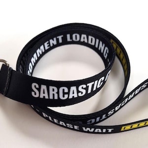 May include: Black lanyard with the words "SARCASTIC COMMENT LOADING PLEASE WAIT" in white text. The lanyard has a silver-coloured metal clasp. The edges of the lanyard have a yellow stripe.