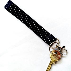 May include: A black fabric wrist strap with white polka dots, connected to a gold-coloured key and a metal clasp. The key is engraved with the word "Timpson".
