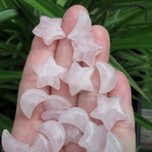 May include: A collection of small, pale pink rose quartz star and crescent moon shaped gemstones. The polished stones are held in a hand, with green foliage in the background. The stars have five points, and the moons are crescent shaped.
