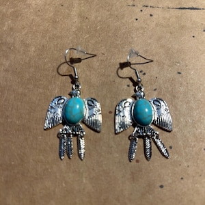 May include: A pair of silver-toned earrings featuring a turquoise-colored oval stone at the center. The earrings are designed with stylized wings and feather-like details, with a hook closure.