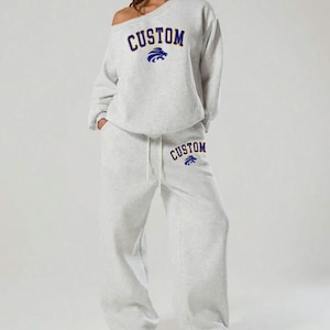 May include: A light gray sweatsuit with the word "CUSTOM" in navy blue arched across the chest and on the left pant leg. A navy blue lion graphic is above the word "CUSTOM". The sweatshirt is off-the-shoulder.