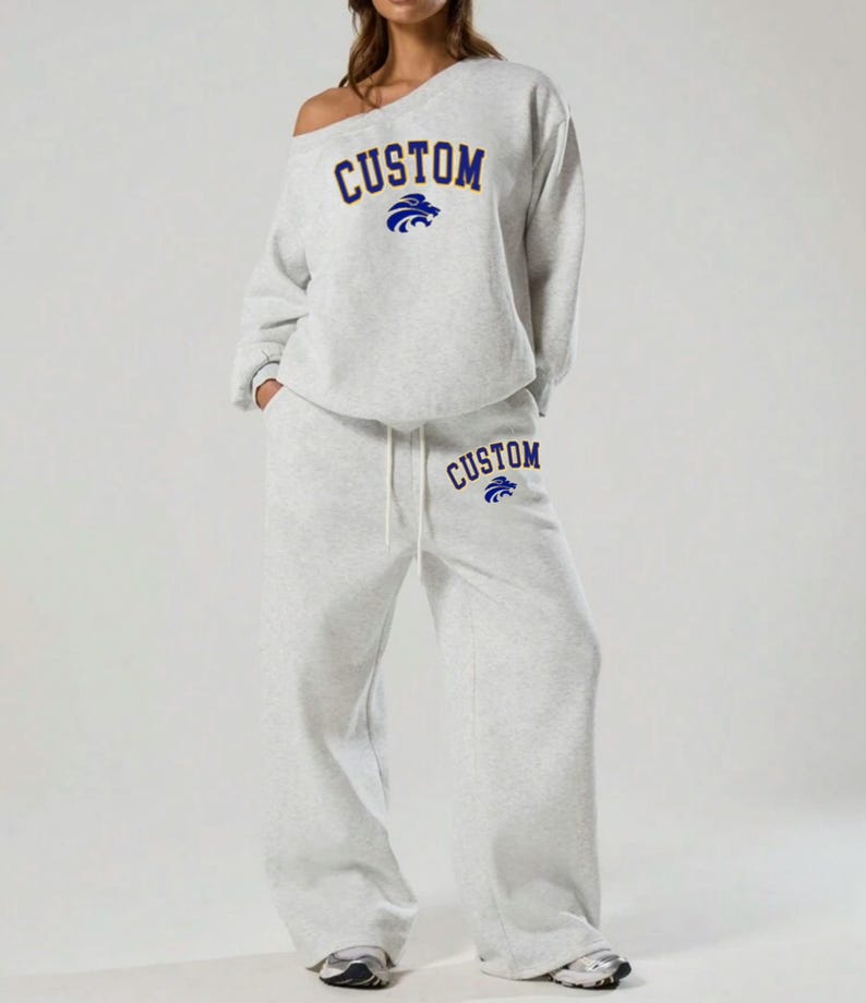 May include: A light gray sweatsuit with the word "CUSTOM" in navy blue arched across the chest and on the left pant leg. A navy blue lion graphic is above the word "CUSTOM". The sweatshirt is off-the-shoulder.