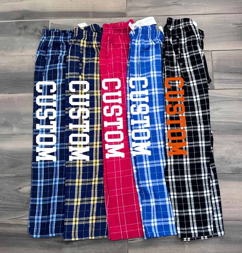 May include: Five pairs of plaid pajama trousers in navy blue, yellow, red, blue, and black. Each pair features the word "CUSTOM" printed vertically in a contrasting colour.